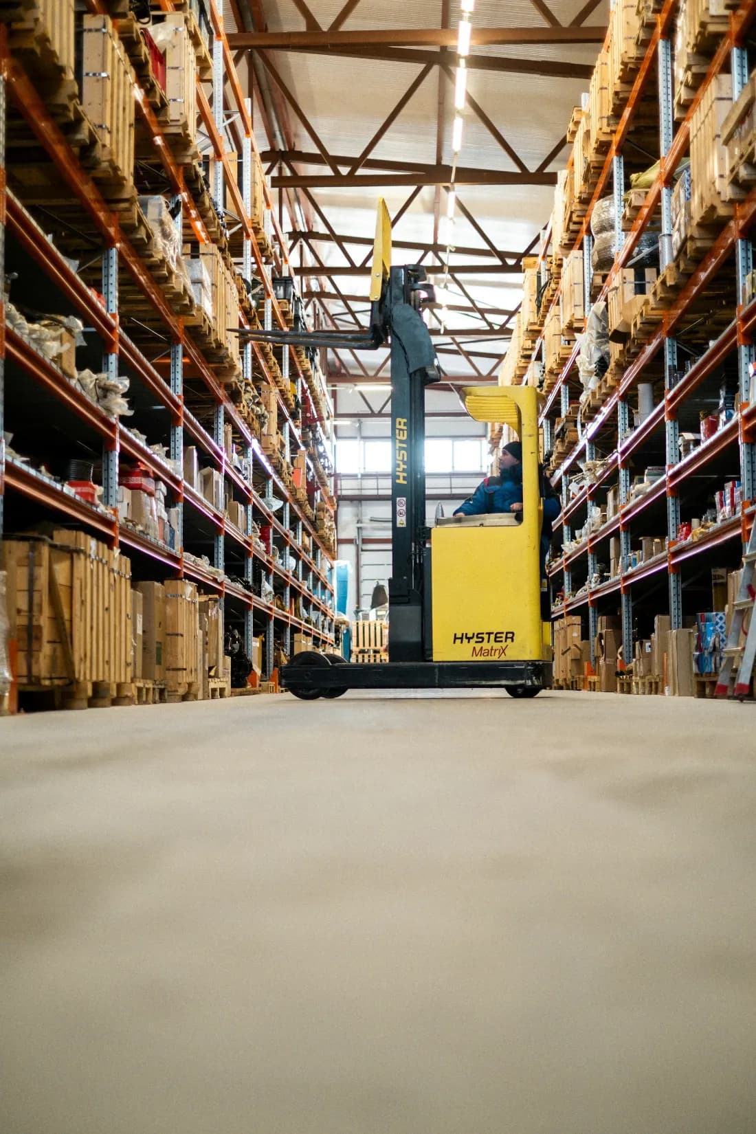 Operator driving a reach truck down a warehouse aisle, racking stacked high with boxed stock on both sides.