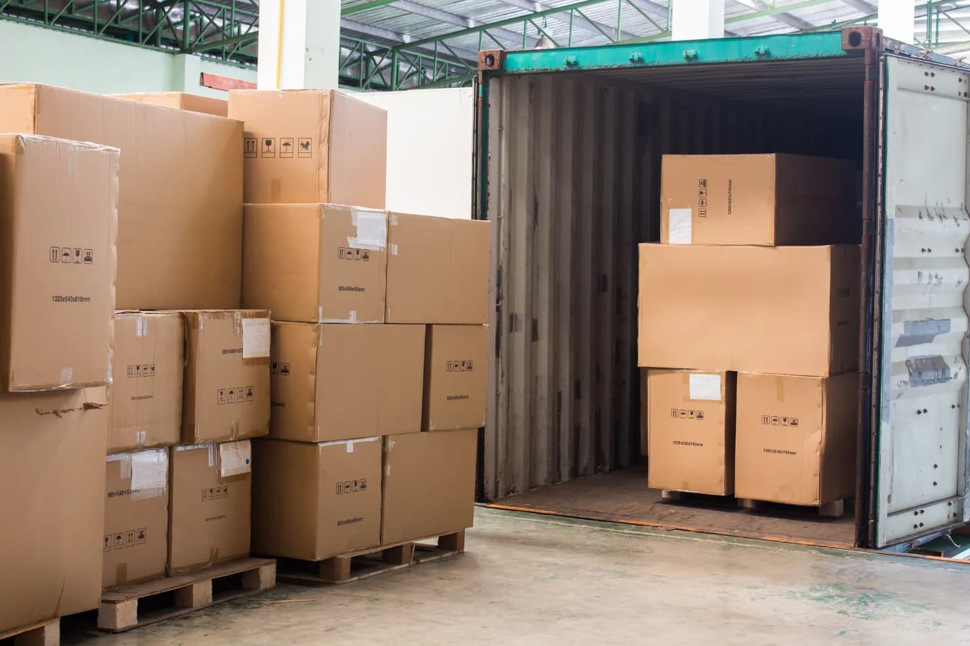 Cases stacked on pallets beside an open shipping container in a warehouse.
