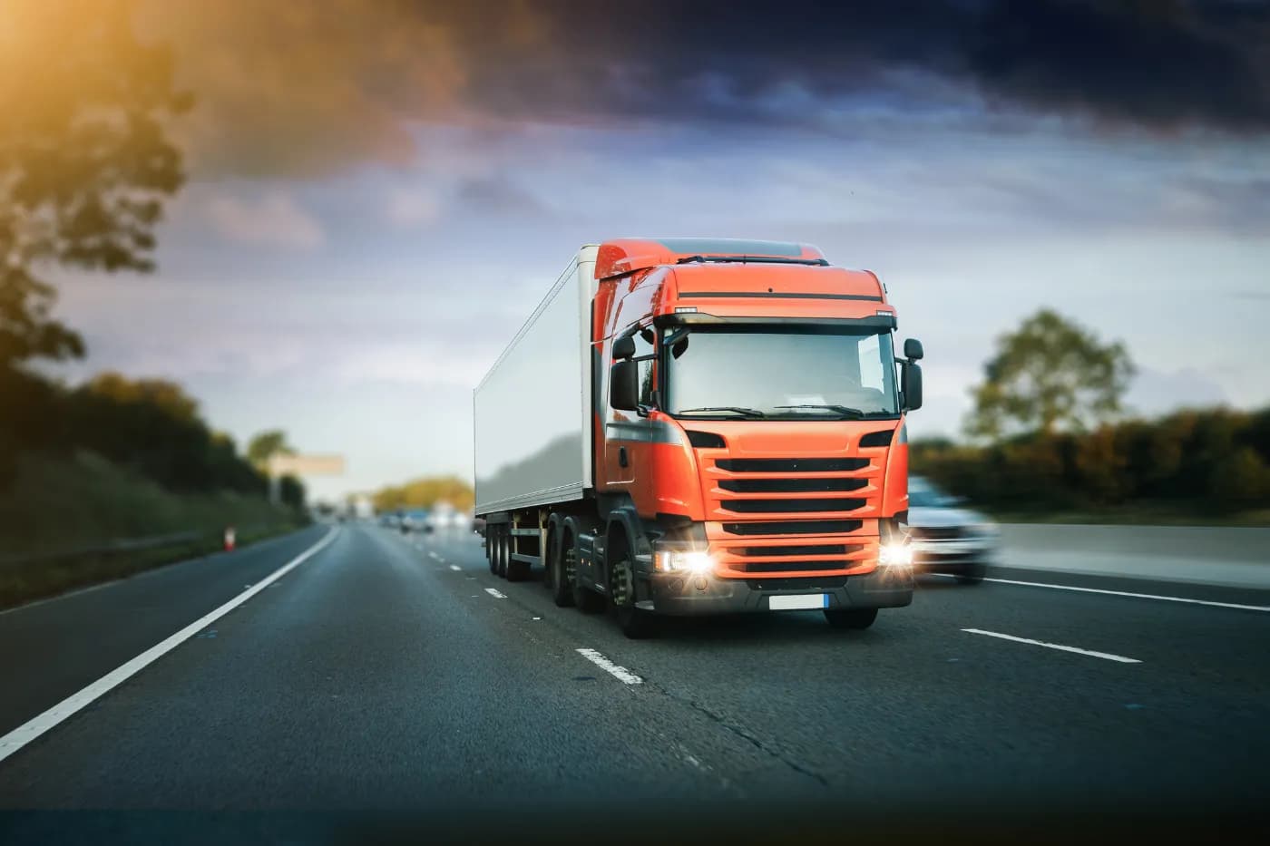 An articulated lorry with an orange cab on a motorway at dusk, headlights on.