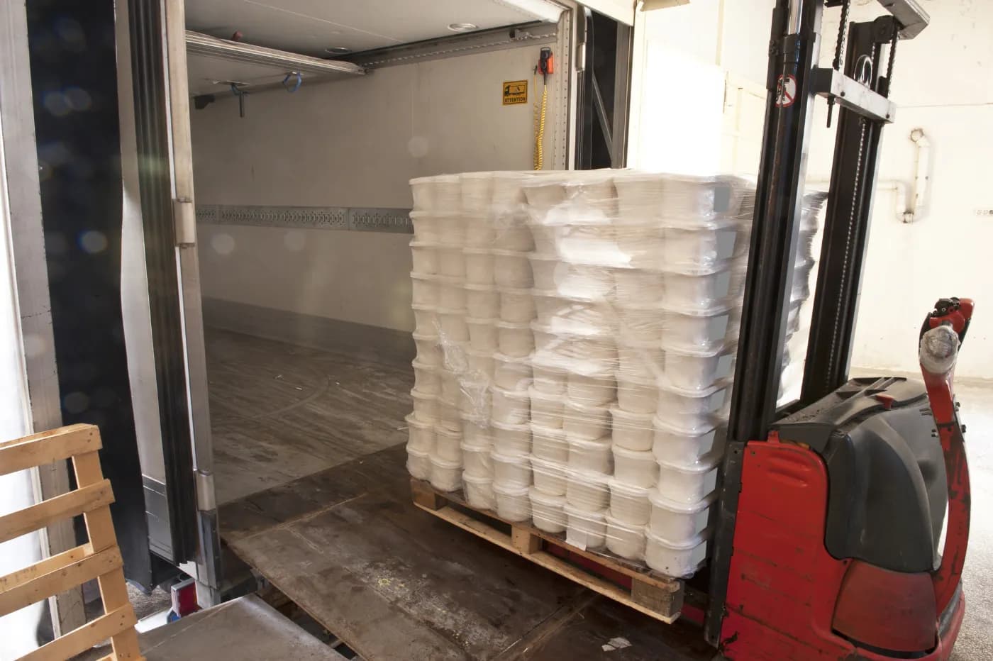 Pallet stacker loading a shrink-wrapped pallet of white tubs into a lorry trailer at a bay.